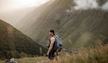 man in gray jacket and black backpack standing on green grass field near mountain during daytime