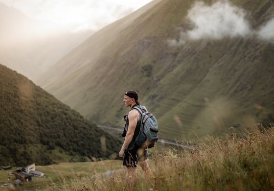 man in gray jacket and black backpack standing on green grass field near mountain during daytime